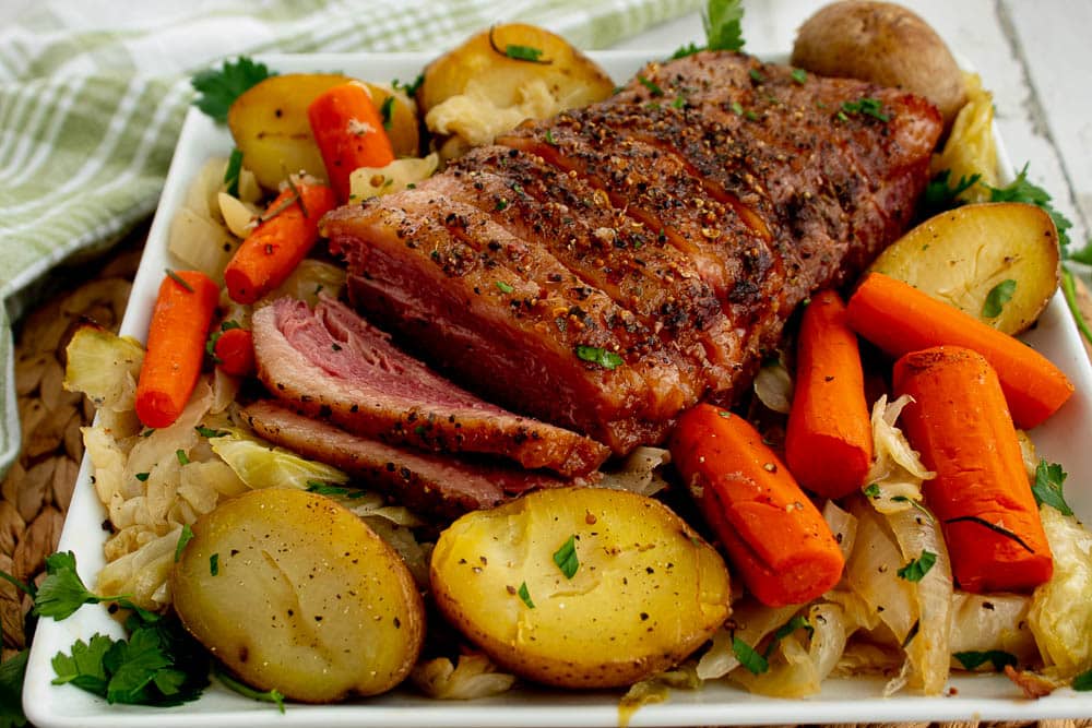 Cooked corned beef with cooked cabbage, onions, carrots, and potatoes on a white platter on a bamboo placemat with a green towel.