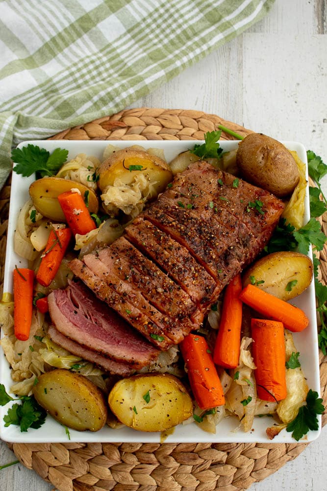 Cooked corned beef with cooked cabbage, onions, carrots, potatoes, and parsley on a white platter on a bamboo placemat with a green towel.