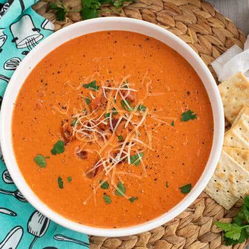 Homemade Bacon Tomato Soup in a white serving bowl on a bamboo placemat, saltines, and blue and white towel.