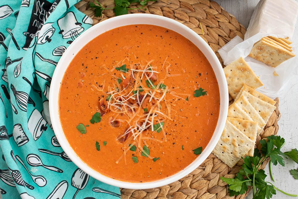 Homemade Bacon Tomato Soup in a white bowl with saltine crackers on a bamboo placemat with a blue and white towel.