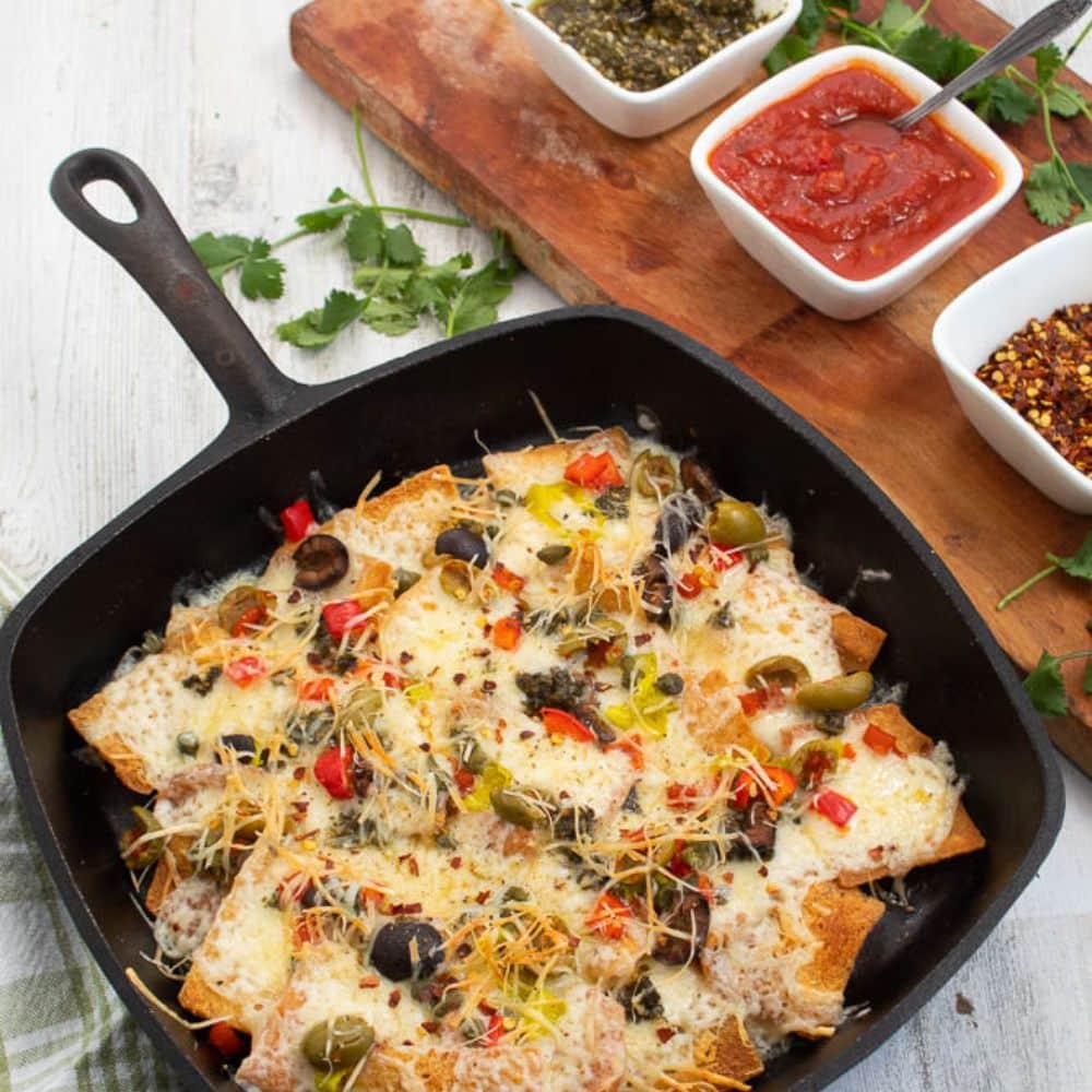 Baked Italian nachos in a black cast iron skillet with pesto, marinaria sauce, and red pepper flakes in white serving dishes on a cutting board.