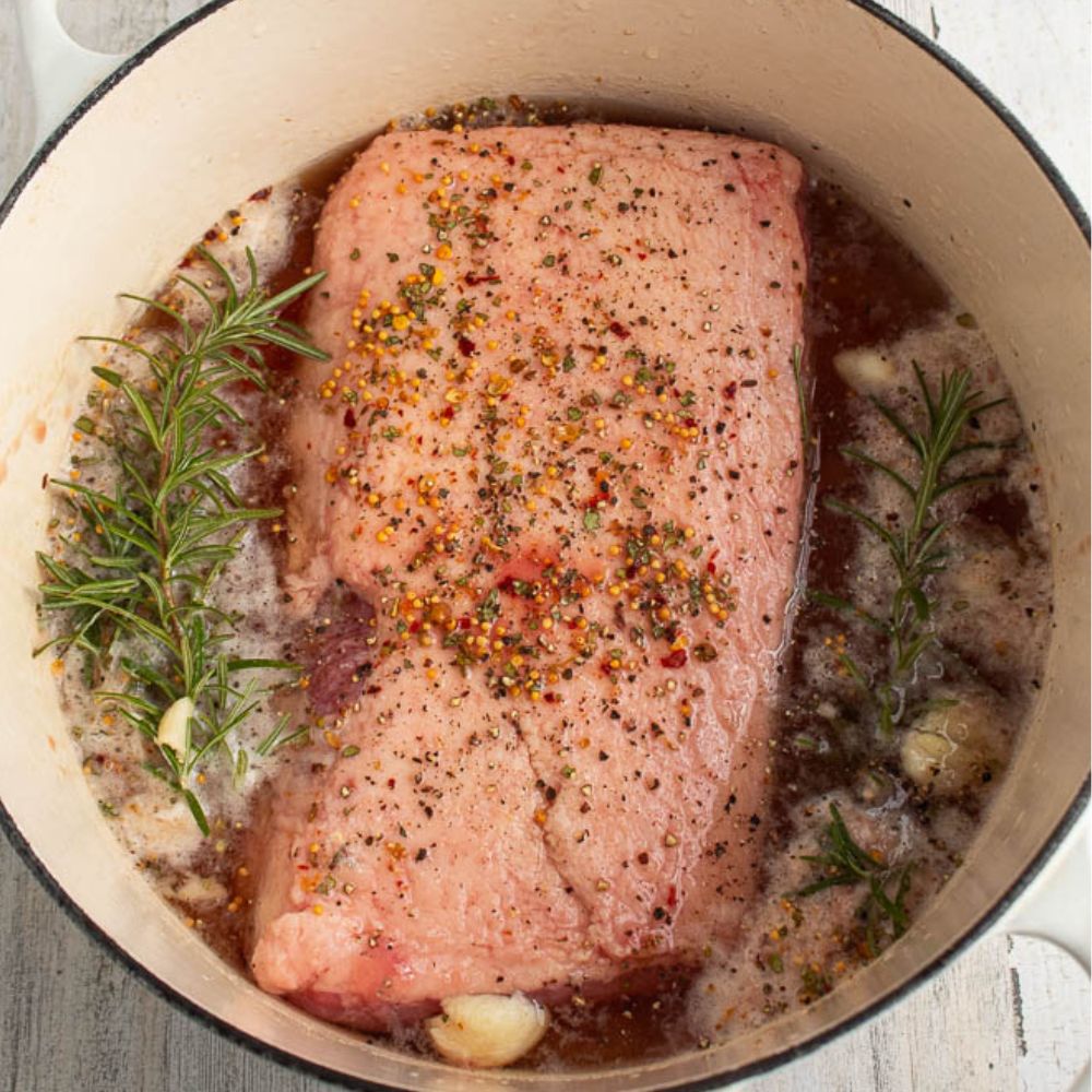 Corned beef in a white Dutch oven with rosemary sprigs, garlic, and seasonings on top of corned beef.