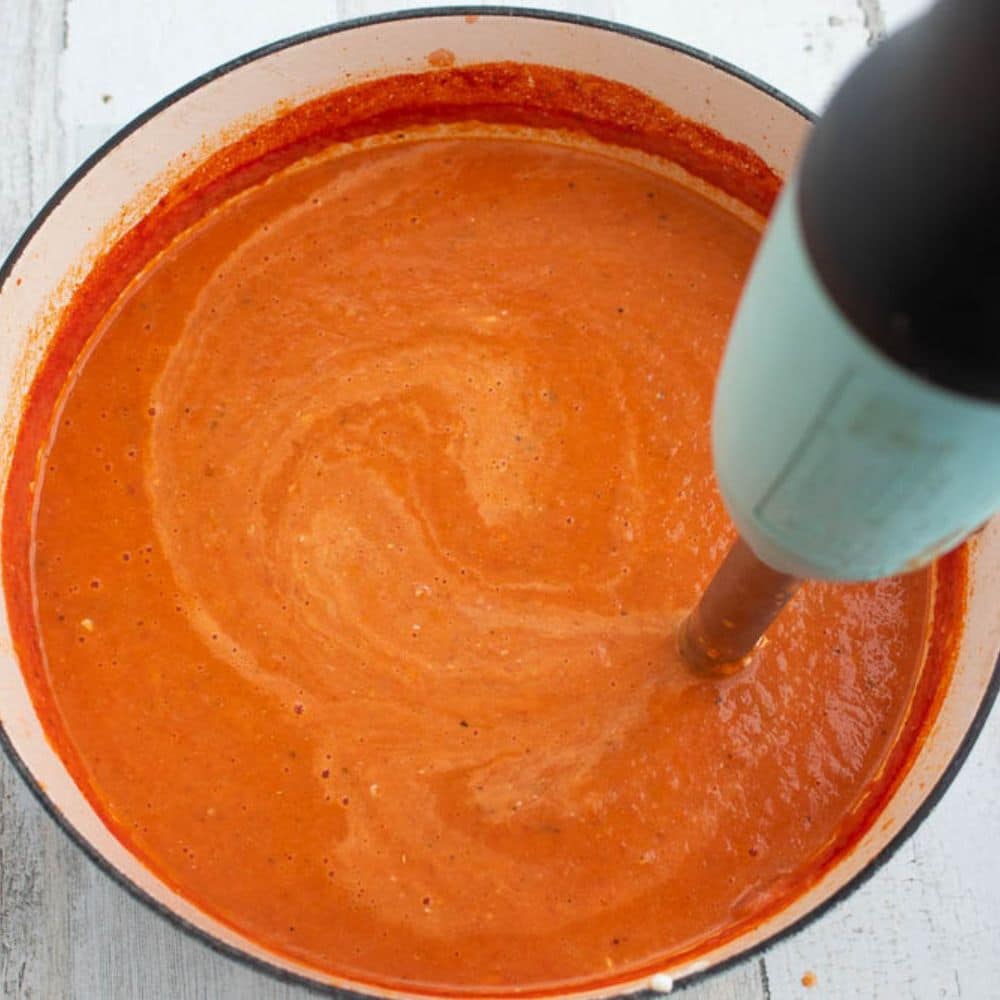 Bacon tomato soup with cream cheese added being blended with an immersion blender in a white Dutch oven.