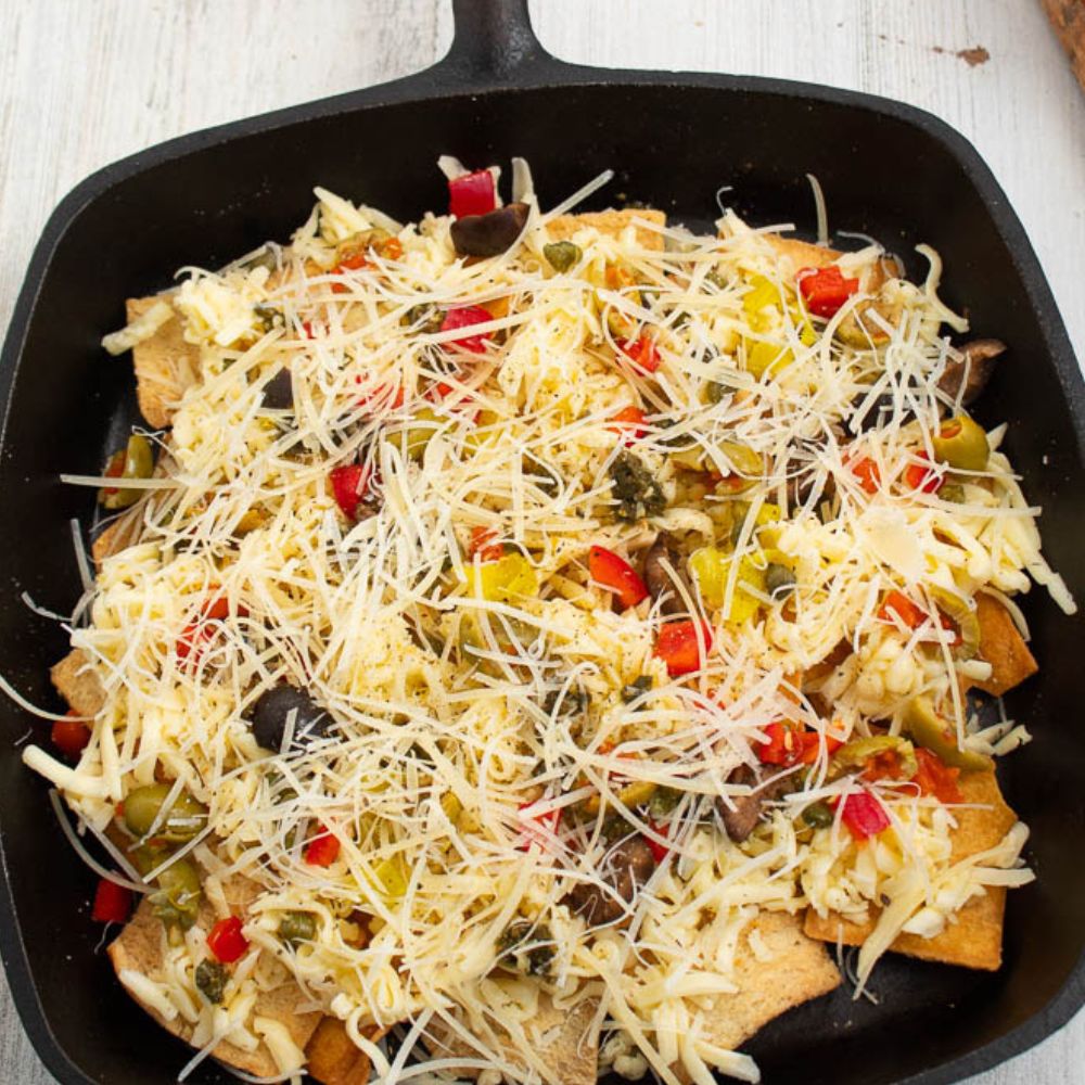 Pita chips in a black cast iron skillet with pesto, grated cheese, olives, red peppers, banana peppers, italian seasoning, and parmasen cheese on top.