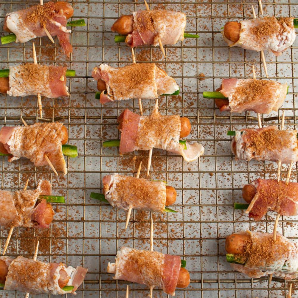 Brown sugar and cinnamon sprinkled on top of uncooked bacon wrapped smokies and jalapeno on a backing rack on top of a cookie sheet.