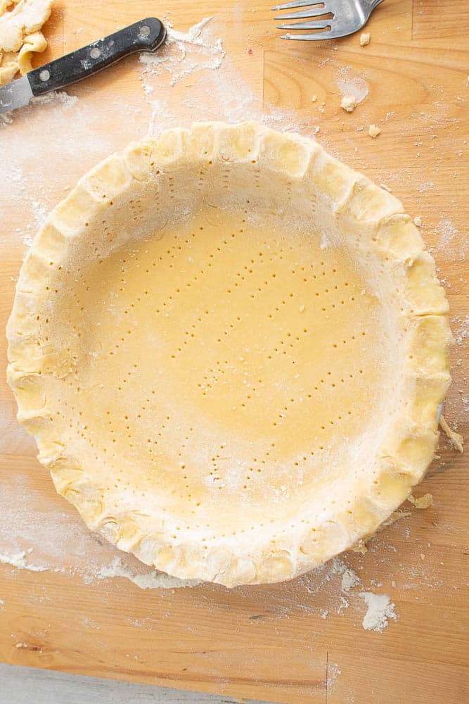 Homemade buttery pie crust in a pie pan with fork holes on a wood cutting board.