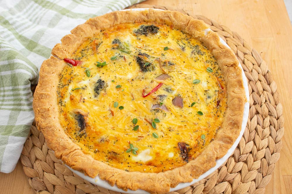 Baked roasted vegetable quiche in a white pie pan, on a bamboo mat with a green and white towel.