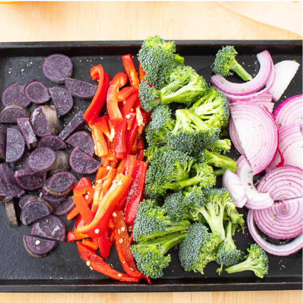 Sliced purple potatoes, red peppers, brocalli, red onions on a baking sheet.