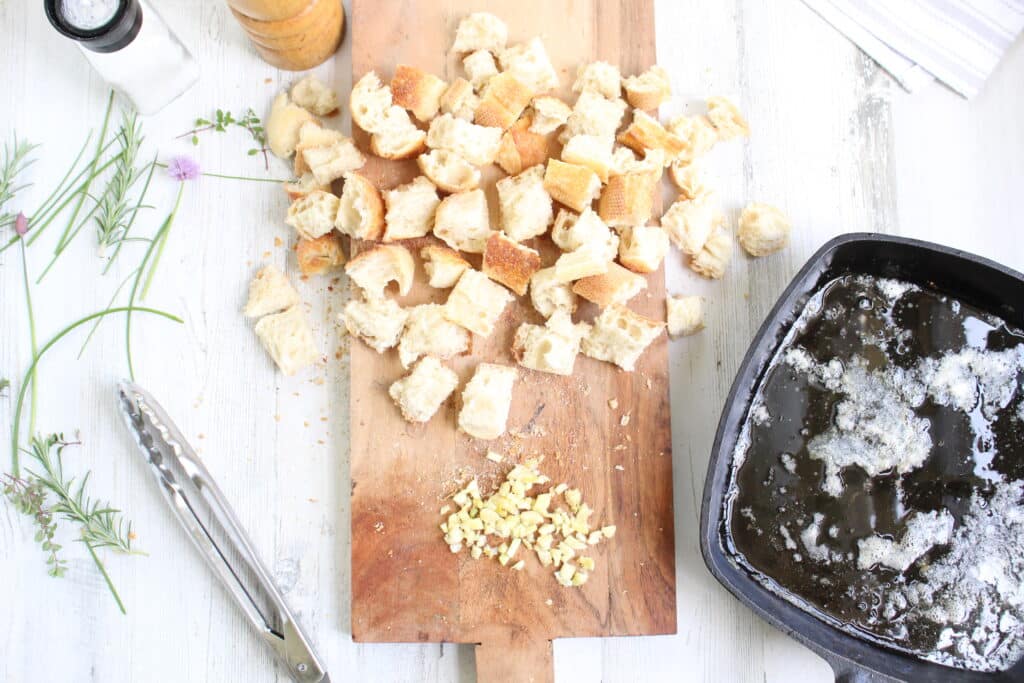 Melted butter in a skillet with bread cubes, garlic, and herbs on a wood cutting board.