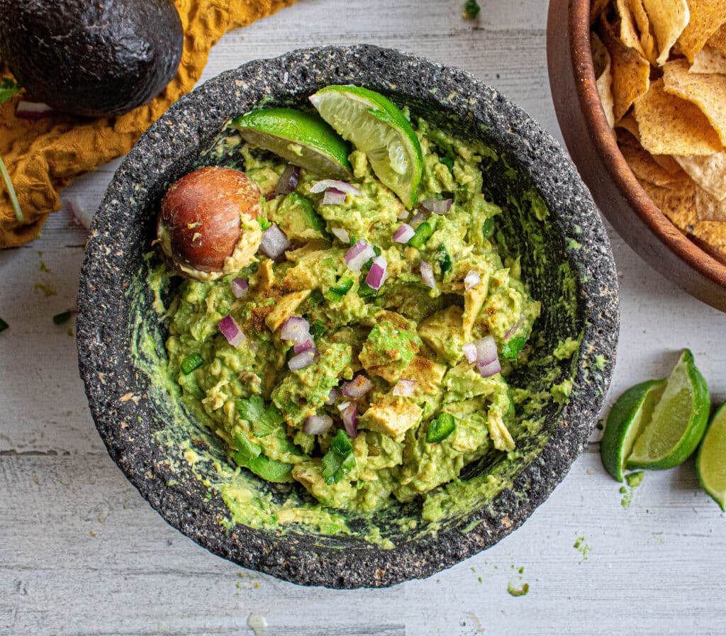 Guacamole in a molcajete bowl with lime, red onion, jalapeno, and seed. Bowl of tortilla chips in a wood bowl with extra avocado..