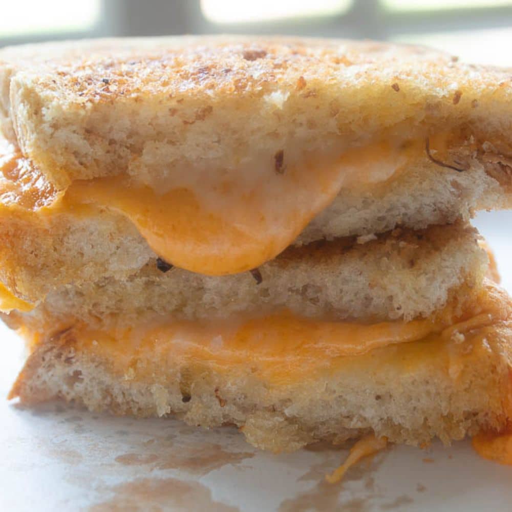 Stack of Grilled Cheese Sandwich Halves on a white paper plate with melted cheese oozing out.