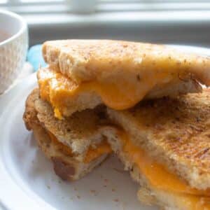 Stack of air fryer grilled cheese sandwich slices on a white paper plate with a bowl of tomato soup.