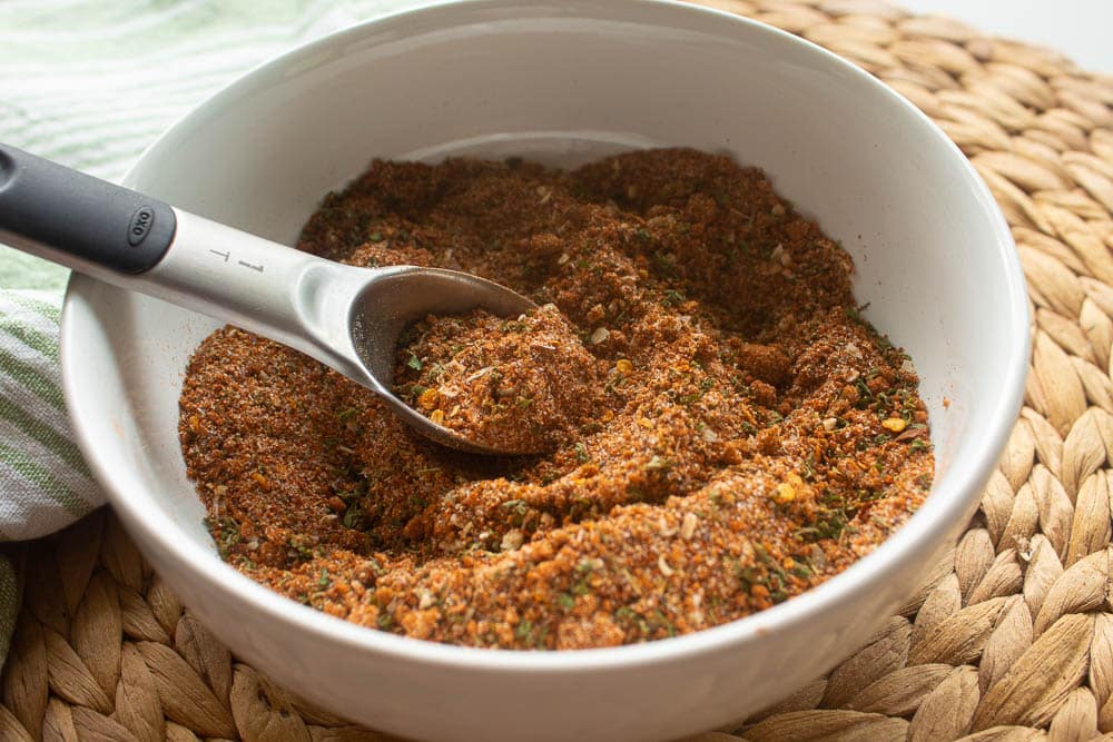 White Bowl full of homemade sweet and spicy jerk seasoning with a tablespoon as a scoop on a tan bamboo placemat with a green and white towel.