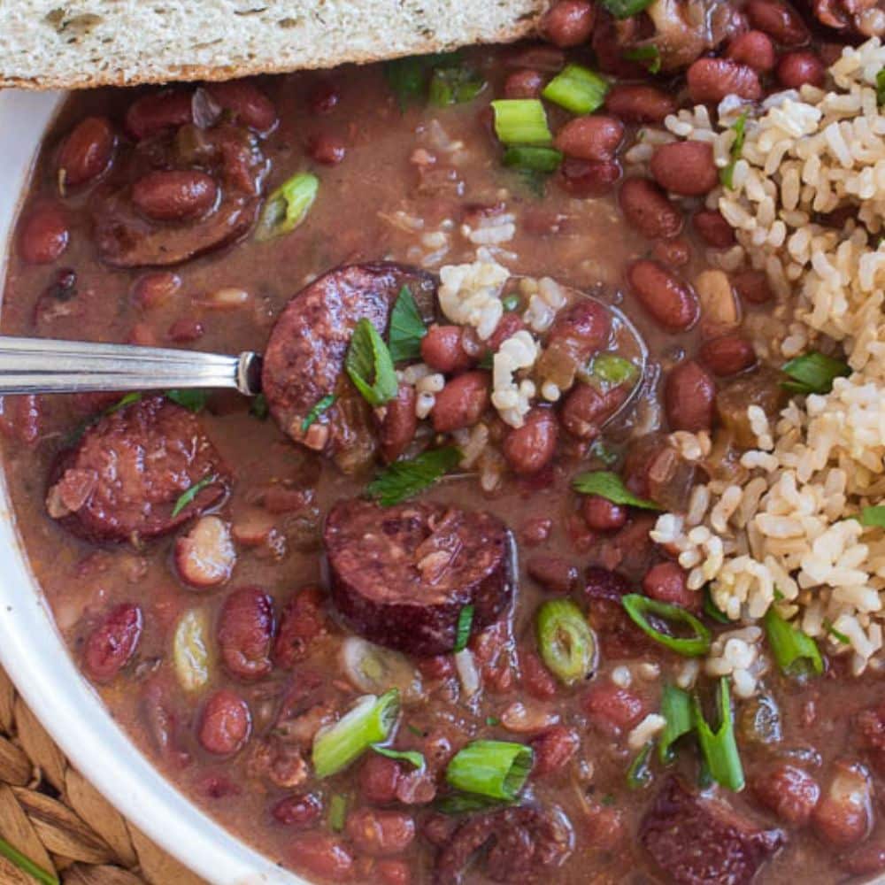 Bowlful of crockpot cajun red beans and rice with chopped green onions and parsly on top in a white bowl with a spoone and piece of french bread.