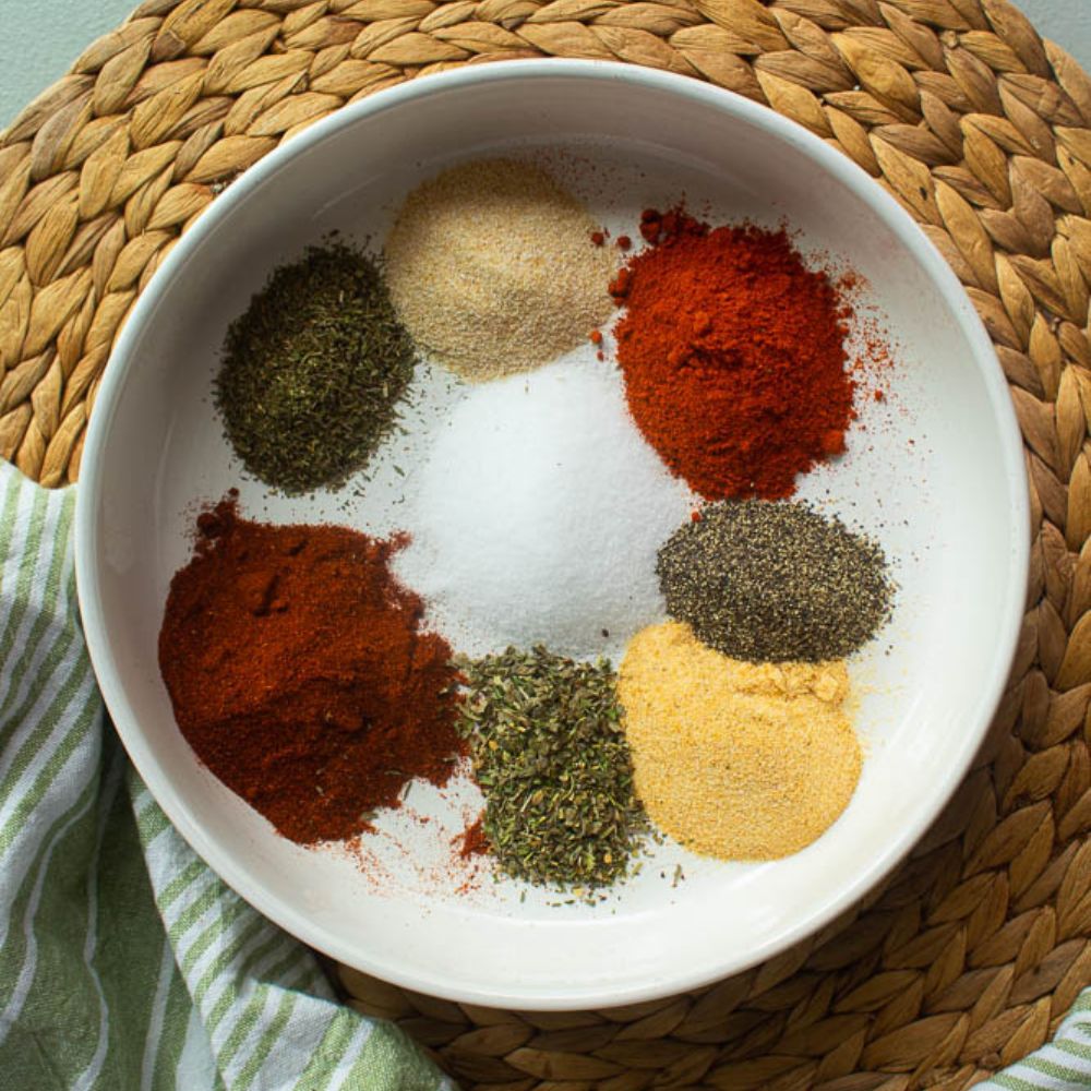 White bowl full of 8 individual different spices to make Cajun seasoning on a tan bamboo mat.