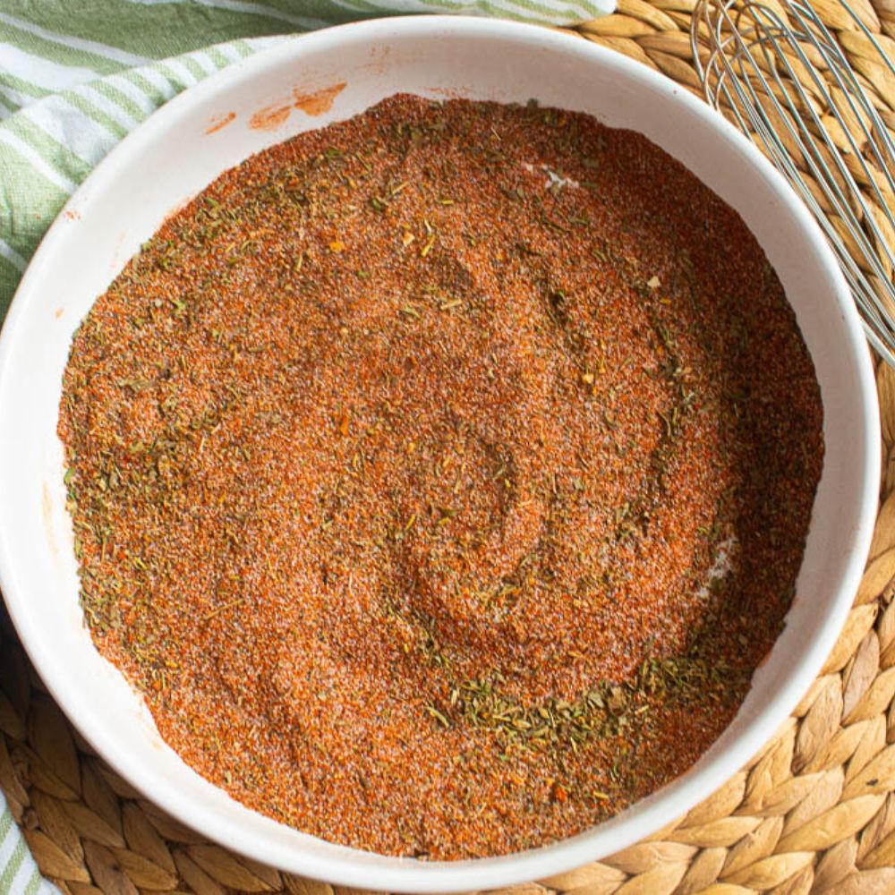 Cajun spice mix in a white bowl on a tan bamboo mat with a whisk.